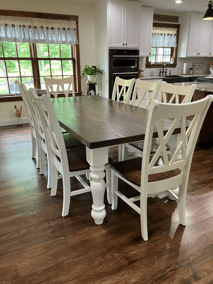 Solid wood farmhouse dining table with dark stained top, white chunky turned legs, and matching white chairs in a bright dining room.